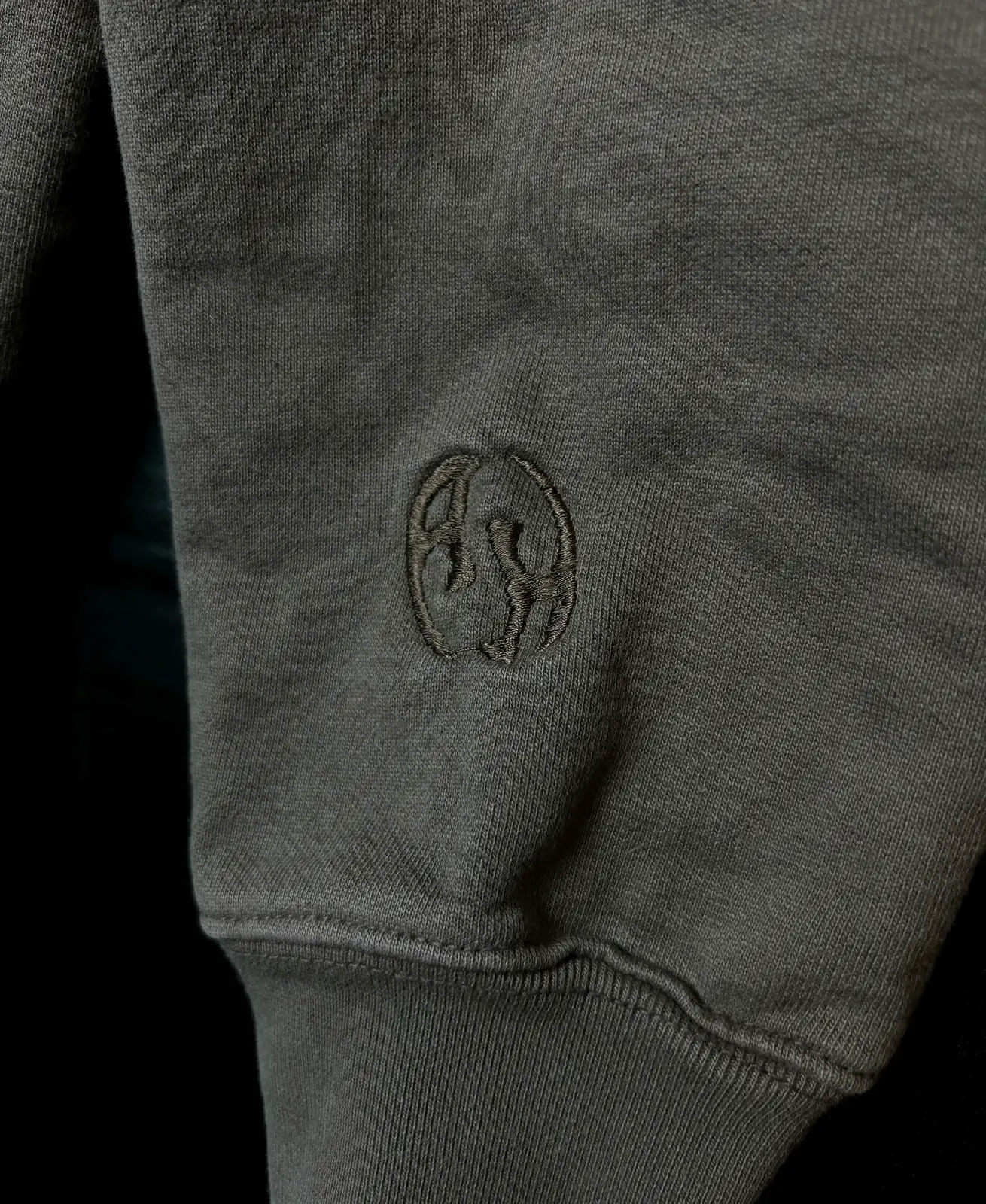 Close-up of a tonal embroidered monogram on the sleeve of a washed olive zip hoodie, just above the cuff